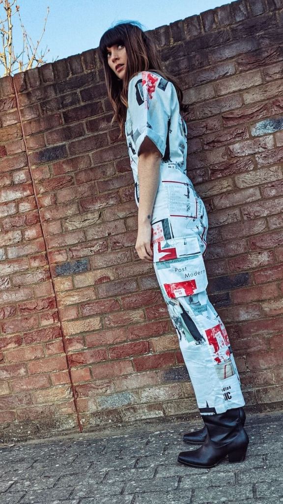 Founder and editor of Wear Next Daisy Jordan wears Fanfare Label printed white boilersuit and black western boots as she stands in front of a red brick wall