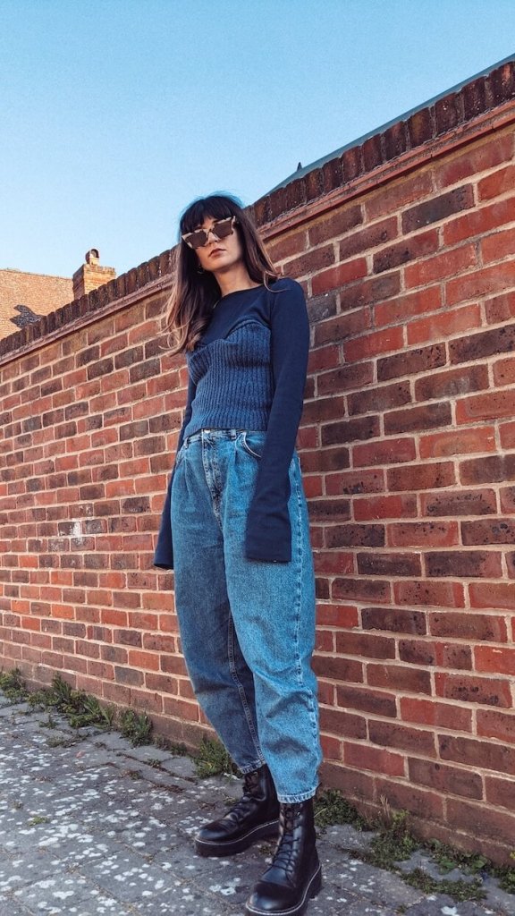 Founder and editor of Wear Next Daisy Jordan wears Fanfare Label navy jumper with bodice, slouchy denim jeans, chunky boots and a pair of tortoiseshell sunglasses as she stands in front of a red brick wall