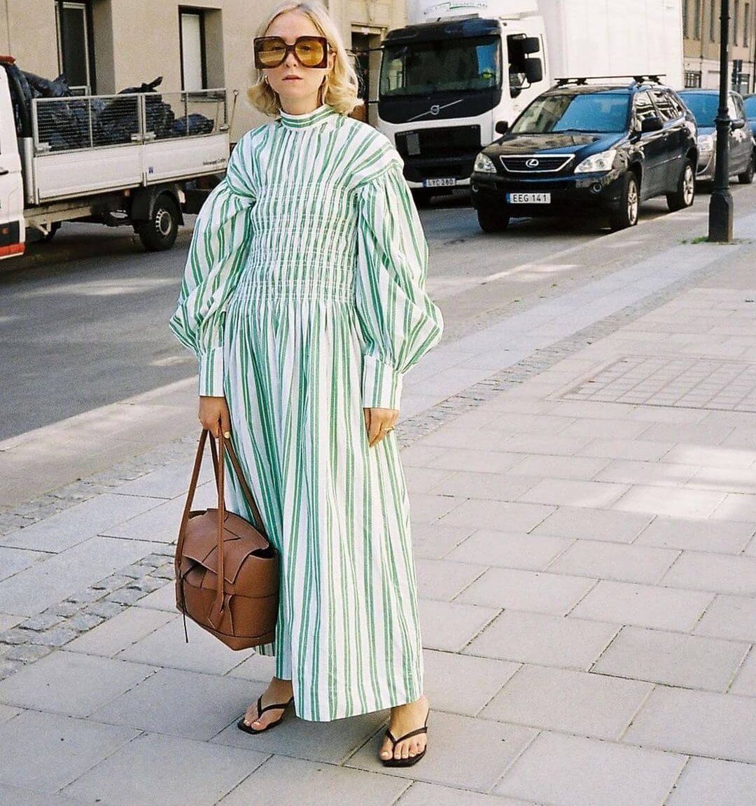 Woman wears oversized 70s sunglasses, a white and green maxi dress with puff sleeves from Ganni, thong sandals and a tan handbag