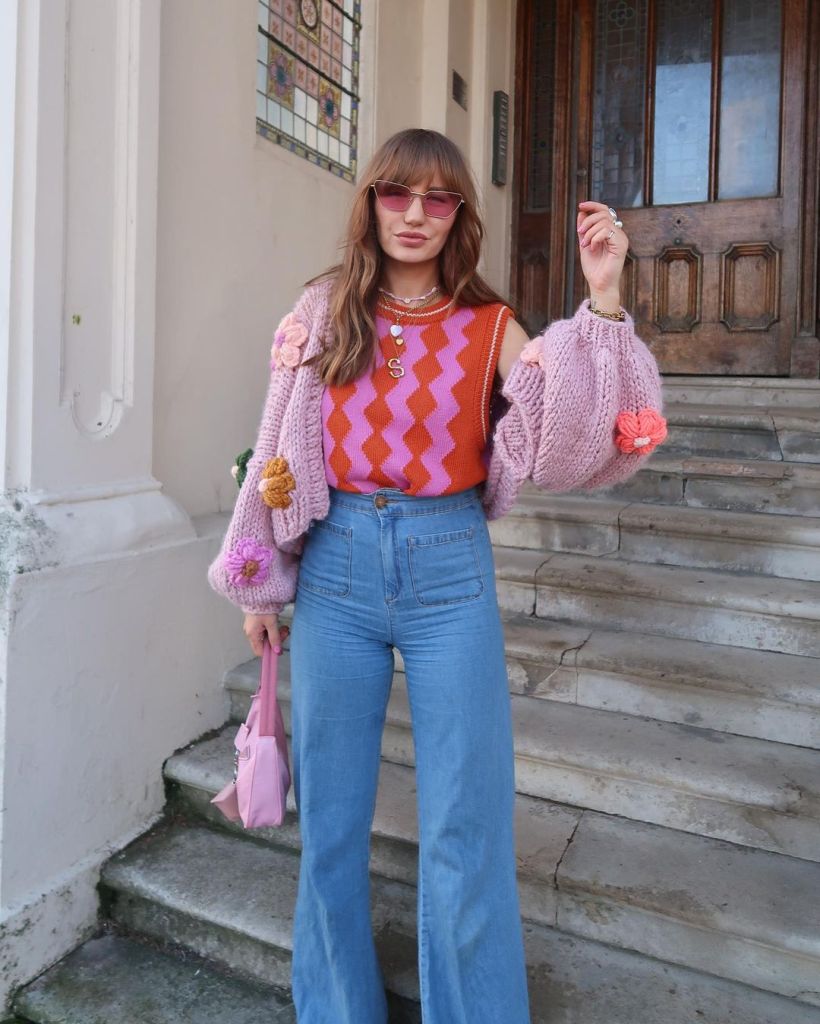 sara waiste wearing a red and pink vest, chunky cardigan and 70s-style wide legged jeans