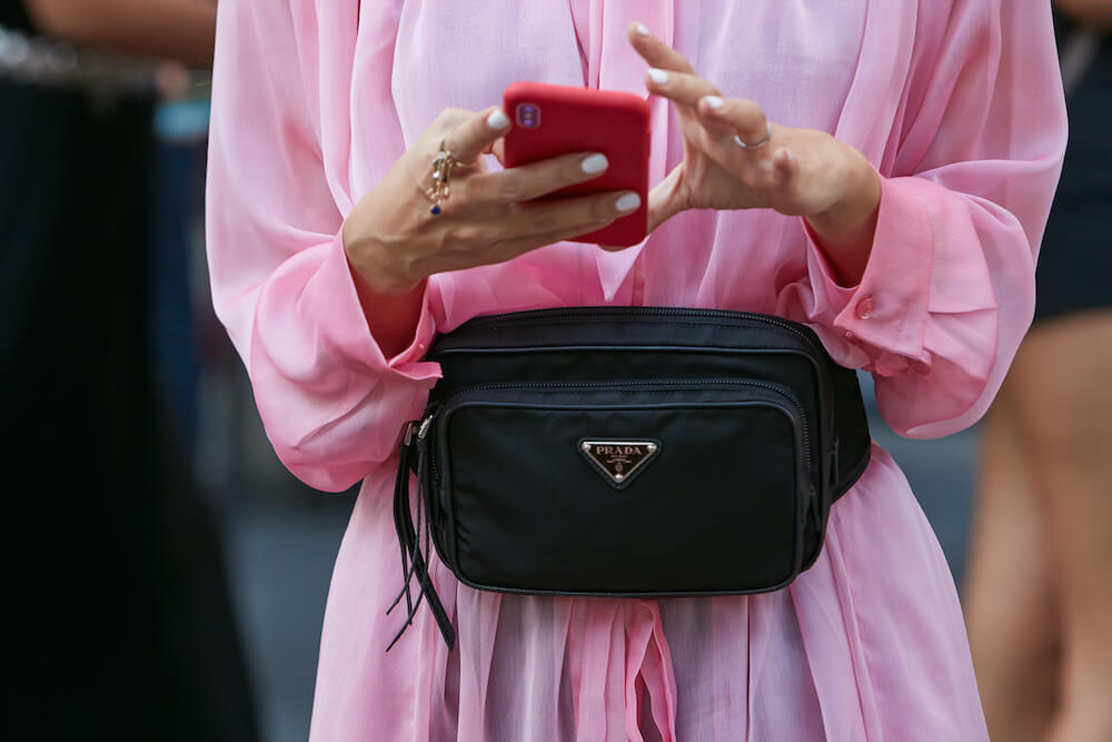 Woman in pink dress holds phone and wears black Prada bum bag