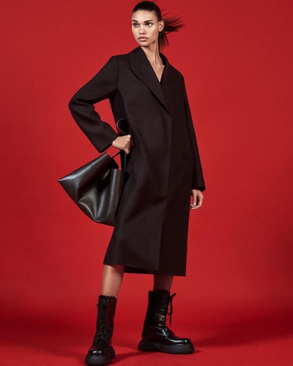 woman wears black coat, leather bag and chunky boots from cos in front of a red background