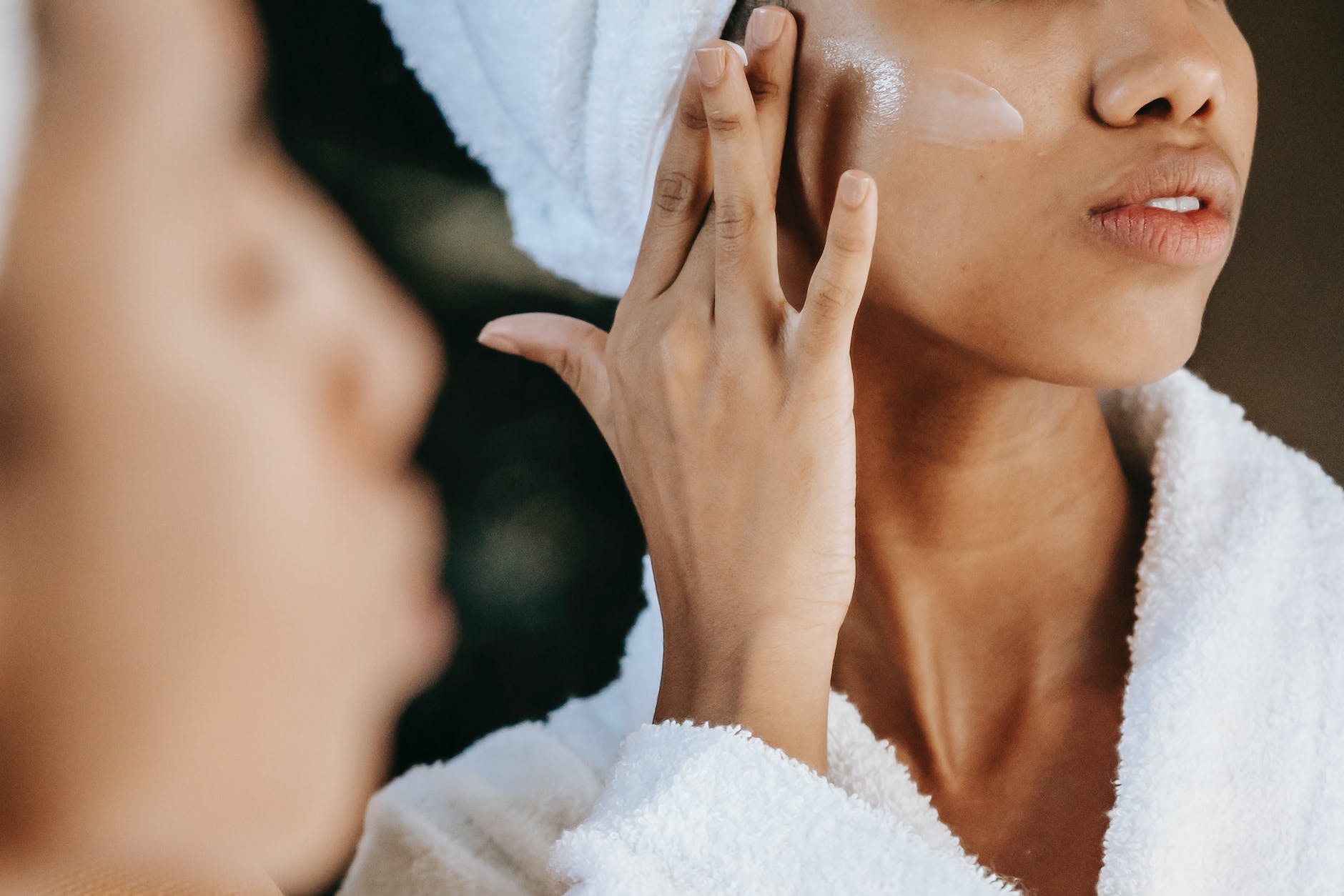 crop ethnic woman applying cream on face against mirror