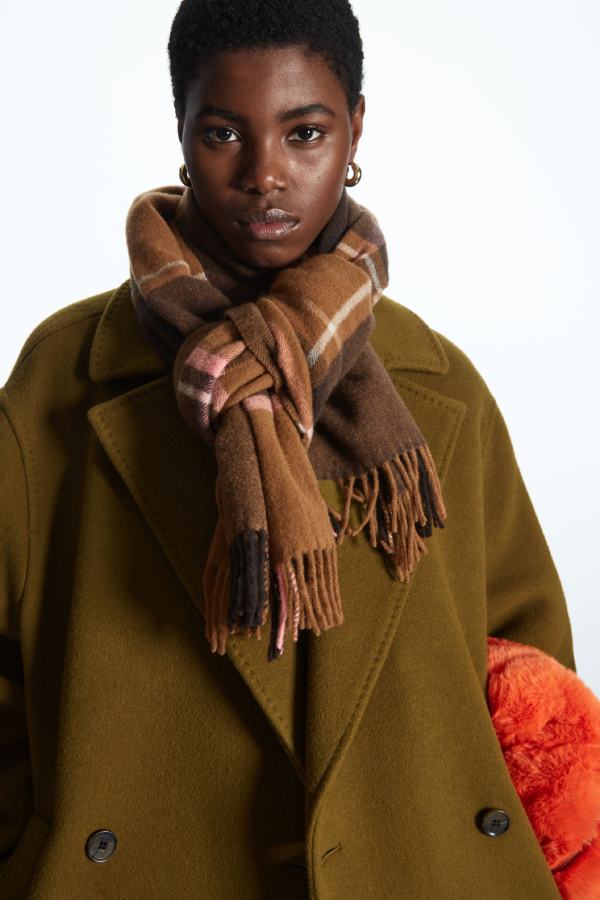Black woman wears olive green coat and a check brown scarf from COS